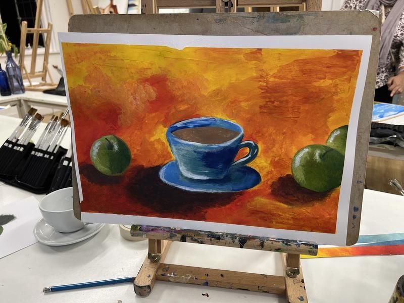 Student acrylic still life of a blue teacup and green apples on an orange background, on an easel
