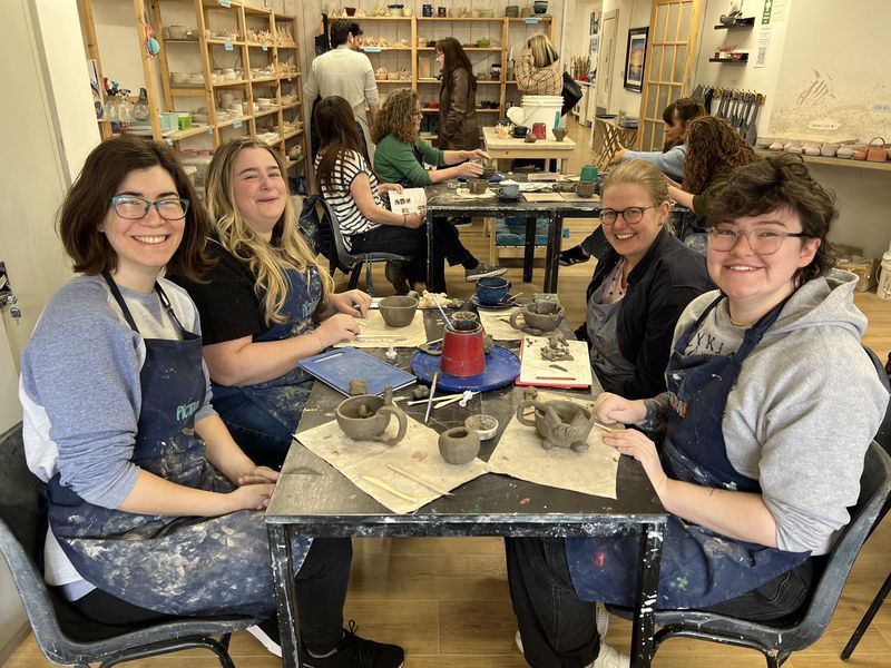 Four smiling adults in aprons around a table of clay at a pottery workshop
