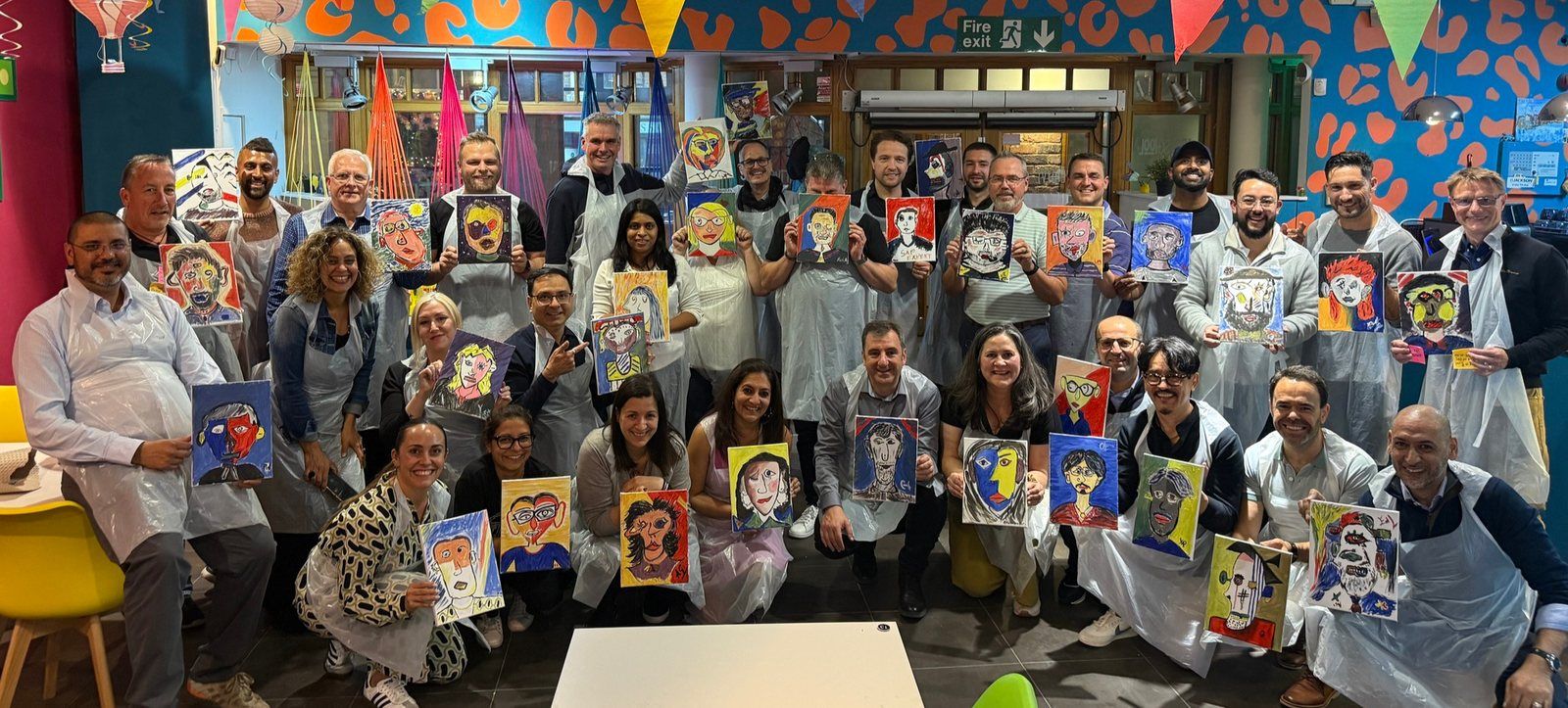 A big corporate group of around 30 colleagues in aprons, each holding up the Picasso-style portrait they painted at their team event